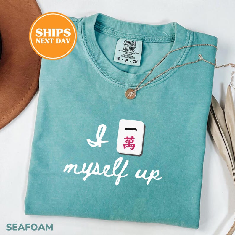 Pu&ograve; includere: T-shirt verde acqua con la scritta bianca "I 10.000 myself up". Una collana d'oro con un ciondolo &egrave; appoggiata sulla maglietta. La maglietta ha uno scollo tondo e una piccola etichetta. La maglietta &egrave; piegata.