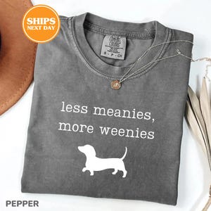 A gray t-shirt with the text "less meanies, more weenies" printed on it, along with a small white dog silhouette. The t-shirt is displayed on a white background, and there is a brown hat and some beige fabric visible in the background.