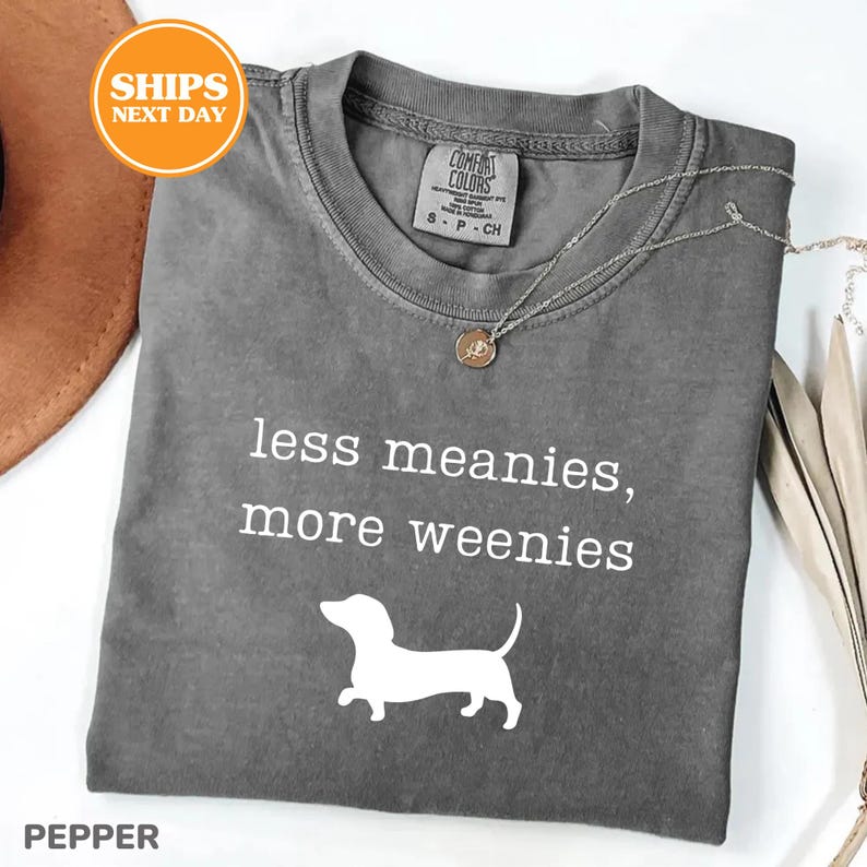 A gray t-shirt with the text "less meanies, more weenies" printed on it, along with a small white dog silhouette. The t-shirt is displayed on a white background, and there is a brown hat and some beige fabric visible in the background.