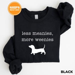A black t-shirt with the text "less meanies, more weenies" printed on it, along with a white silhouette of a dachshund dog.