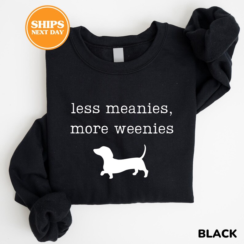 A black t-shirt with the text "less meanies, more weenies" printed on it, along with a white silhouette of a dachshund dog.