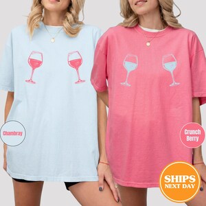 May include: Two short-sleeved t-shirts, one light blue and one pink, each featuring a wine glass graphic. The light blue shirt has a red wine glass design, while the pink shirt has a blue wine glass design. The shirts are casual and comfortable.