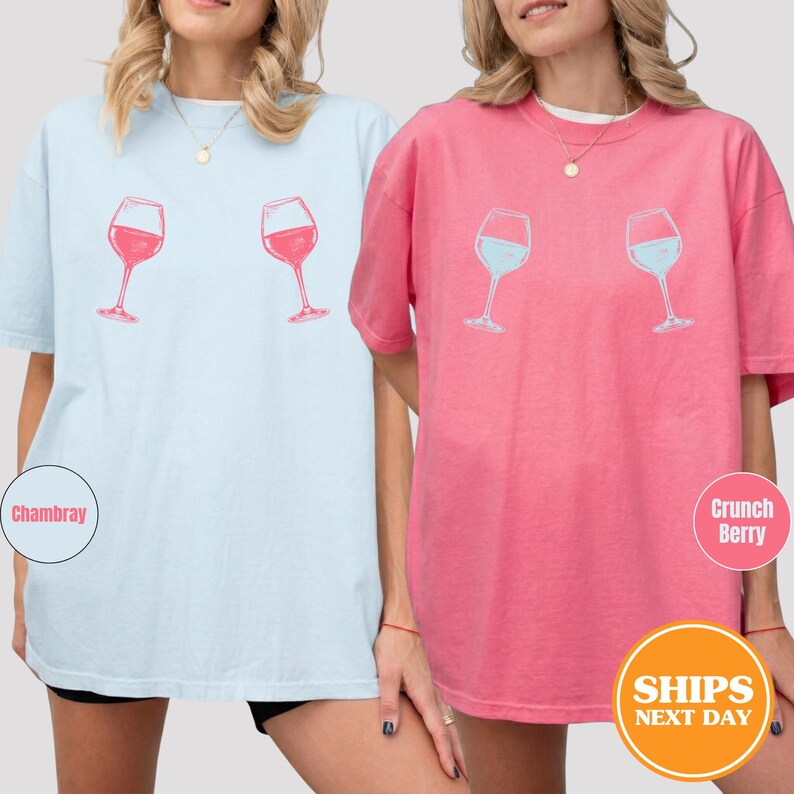 May include: Two short-sleeved t-shirts, one light blue and one pink, each featuring a wine glass graphic. The light blue shirt has a red wine glass design, while the pink shirt has a blue wine glass design. The shirts are casual and comfortable.
