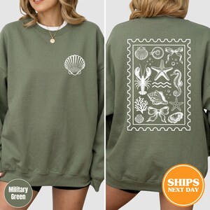 May include: An olive green crewneck sweatshirt featuring a white line art graphic of a single seashell on the front left chest. The back displays a large rectangular stamp-shaped design containing various marine life illustrations including a lobster, starfish, seahorse, shells, coral, and two decorative bows.