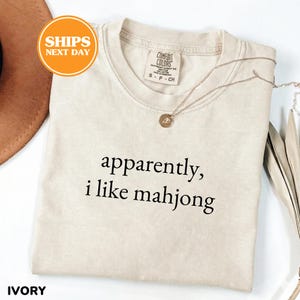 May include: Ivory-colored t-shirt with the text "apparently, i like mahjong" in black. The shirt has a crew neck and short sleeves. A gold necklace with a pendant is draped across the shirt. The shirt is labeled "Comfort Colors".