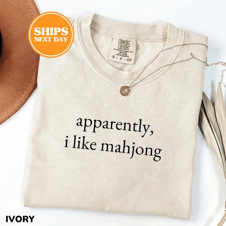 May include: Ivory-colored t-shirt with the text "apparently, i like mahjong" in black. The shirt has a crew neck and short sleeves. A gold necklace with a pendant is draped across the shirt. The shirt is labeled "Comfort Colors".