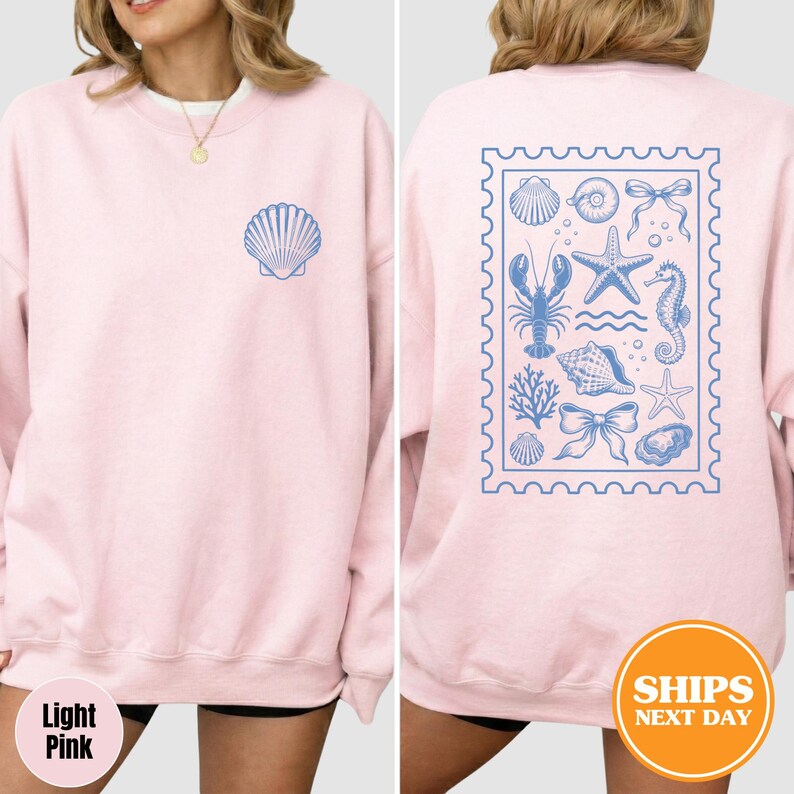 May include: A light pink crewneck sweatshirt featuring a small blue seashell graphic on the front left chest. The back displays a large rectangular stamp-shaped design containing blue illustrations of a lobster, starfish, seahorse, and bows.