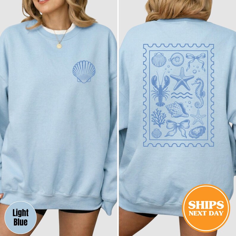 May include: A light blue crewneck sweatshirt featuring a small shell graphic on the front left chest and a large rectangular stamp-shaped design on the back. The back graphic contains illustrations of a lobster, starfish, seahorse, and bows.