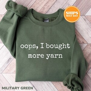 May include: Military green sweatshirt with the text "oops, I bought more yarn" in white. The sweatshirt has a crew neck and long sleeves with ribbed cuffs. The image also includes a "SHIPS NEXT DAY" sticker.