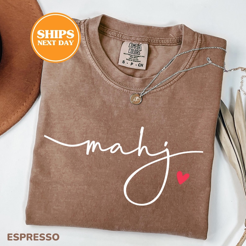 May include: Espresso-colored t-shirt with the word "mahj" in white script and a small red heart. The shirt has a crew neck and a Comfort Colors tag. A silver necklace with a pendant is draped across the shirt.