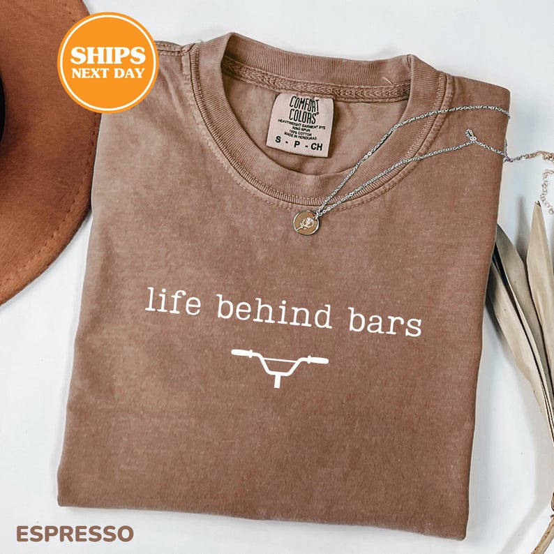 a brown t-shirt with the text "life behind bars" printed on it, along with a bicycle graphic. The t-shirt is laid out on a white surface, and there is a necklace and a brown hat visible in the background.