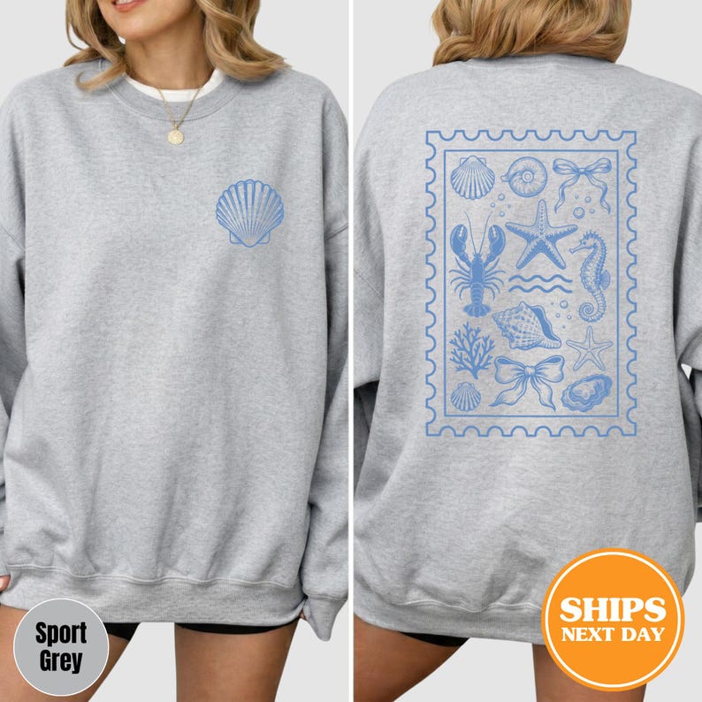 May include: A light gray crewneck sweatshirt featuring a small blue seashell graphic on the front left chest and a large rectangular stamp-style design on the back containing various blue illustrations of sea life, including a lobster, starfish, and seahorse.