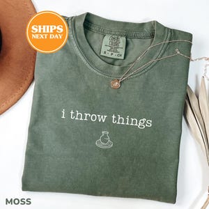 May include: A moss green short-sleeve t-shirt featuring the white text "i throw things" above a small white line drawing of a pottery vase on a wheel. The shirt has a crew neckline and a Comfort Colors brand label printed on the inside collar.