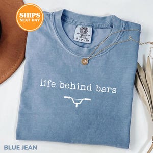 a blue t-shirt with the text "life behind bars" printed on it, along with a bicycle graphic. The t-shirt is laid out on a white surface, and there is a brown hat and some leaves in the background.