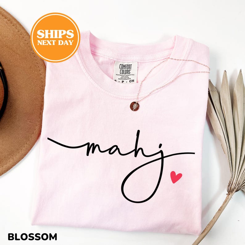 May include: A light pink t-shirt with the word "mahj" in black script and a small red heart. The shirt has a crew neck and is labeled "Blossom". A gold-colored necklace with a pendant is draped on the shirt.