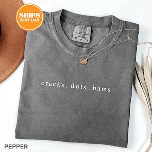 A gray t-shirt with the text "cracks, dots, bams" printed on it, along with a small pendant necklace. The t-shirt is placed on a white surface, and there is a brown hat and some leaves in the background.