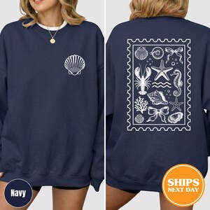 May include: A navy blue crewneck sweatshirt featuring a white shell graphic on the front left chest. The back displays a large white rectangular stamp-shaped design containing various marine life illustrations including a lobster, starfish, and seahorse.