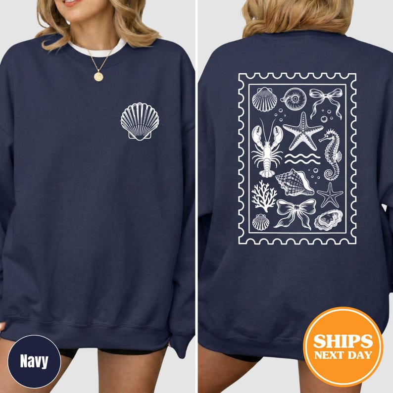 May include: A navy blue crewneck sweatshirt featuring a white shell graphic on the front left chest. The back displays a large white rectangular stamp-shaped design containing various marine life illustrations including a lobster, starfish, and seahorse.