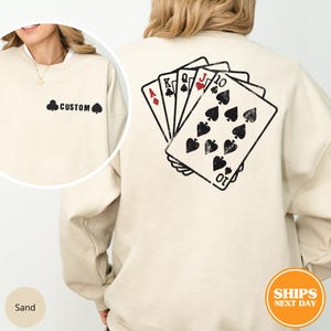 May include: Sand-colored sweatshirt with a black spade symbol and the word "CUSTOM" on the front. The back features a graphic of playing cards: Ace of Diamonds, King of Clubs, Queen of Spades, Jack of Hearts, and Ten of Spades.