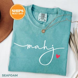 May include: Seafoam green t-shirt with the word "mahj" in white script and a small red heart. A gold necklace with a pendant rests on the shirt. The shirt is folded and has a tag that says "Comfort Colors".