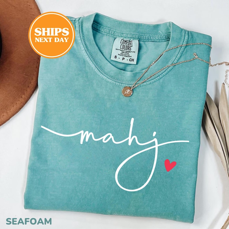 May include: Seafoam green t-shirt with the word "mahj" in white script and a small red heart. A gold necklace with a pendant rests on the shirt. The shirt is folded and has a tag that says "Comfort Colors".