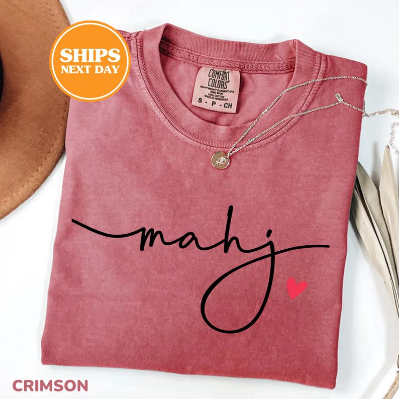 May include: Crimson-colored t-shirt with the word "mahj" in a black script font and a small red heart. A silver necklace with a pendant is draped on the shirt. The shirt is labeled "Comfort Colors" and is a size small.