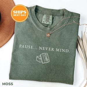 May include: A moss green short-sleeve t-shirt featuring the white text PAUSE... NEVER MIND. and a white line drawing of two mahjong tiles. The shirt has a crew neckline and a Comfort Colors brand label printed on the inside of the collar area.