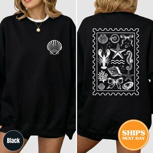 May include: A black long-sleeve sweatshirt featuring a white graphic of a single seashell on the front left chest. The back displays a large white rectangular stamp-shaped design containing illustrations of a lobster, starfish, and shells.