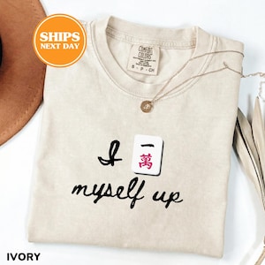 Pu&ograve; includere: T-shirt color avorio con la frase "I myself up" e una grafica a piastrella Mahjong. La maglietta ha girocollo e maniche corte. Una collana d'oro con un ciondolo &egrave; drappeggiata sulla maglietta.