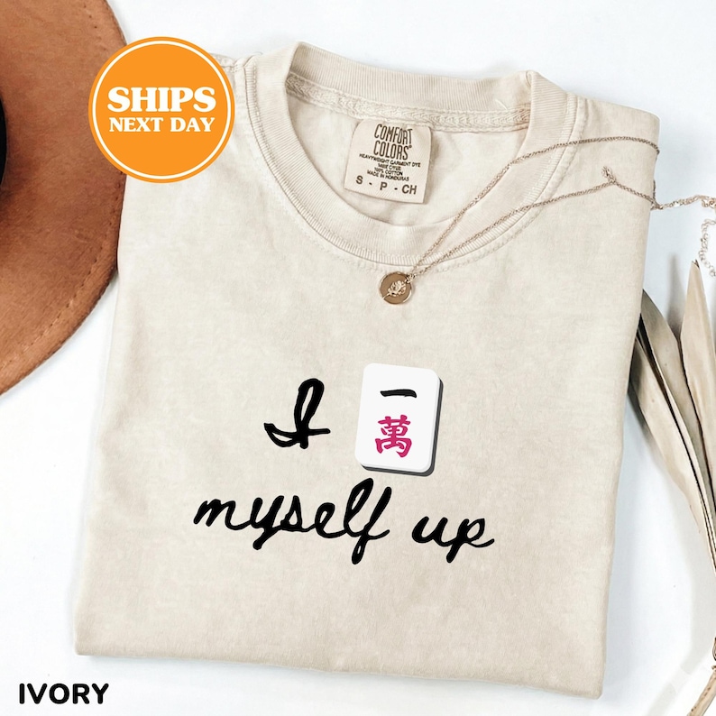 Pu&ograve; includere: T-shirt color avorio con la frase "I myself up" e una grafica a piastrella Mahjong. La maglietta ha girocollo e maniche corte. Una collana d'oro con un ciondolo &egrave; drappeggiata sulla maglietta.