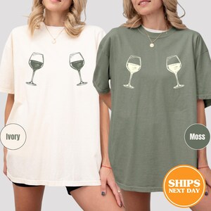 May include: Two short-sleeved t-shirts, one ivory and one moss green, each featuring a sketch of two wine glasses. The ivory shirt has glasses with a dark fill, while the moss shirt has glasses with a light fill. The shirts are casual and comfortable.