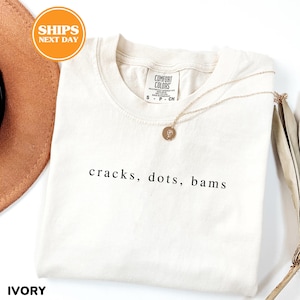 a white t-shirt with the text "cracks, dots, bams" printed on it, along with a necklace and a brown hat.