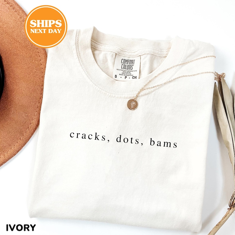 a white t-shirt with the text "cracks, dots, bams" printed on it, along with a necklace and a brown hat.