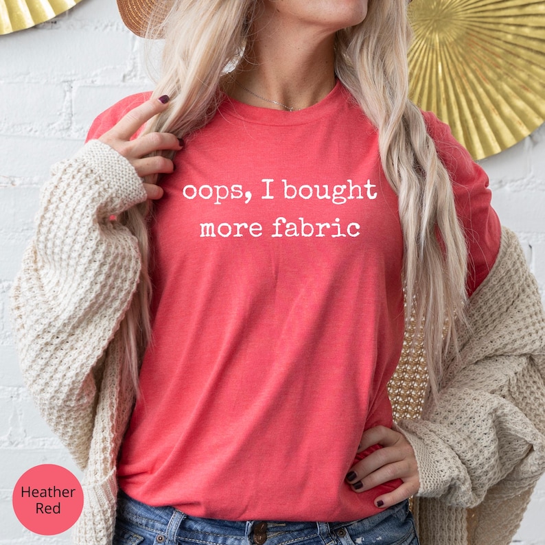A woman wearing a red t-shirt with the text "oops, I bought more fabric" printed on it, standing in front of a white brick wall with a gold fan decoration.