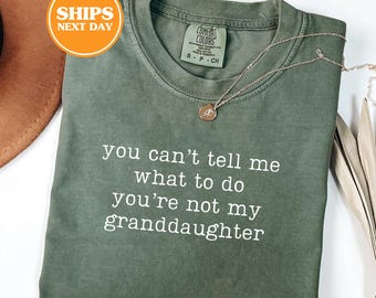 May include: A moss green short-sleeve t-shirt featuring the white printed text, you can't tell me what to do you're not my granddaughter. The shirt has a crew neckline and a Comfort Colors brand label on the inside collar, paired with a gold necklace.