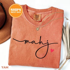 May include: Coral-colored t-shirt with the word "mahj" in a black script font and a small red heart. A gold necklace with a pendant rests on the shirt. The shirt has a Comfort Colors tag.