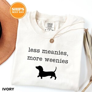 A white t-shirt with the text "less meanies, more weenies" printed on it, along with a black silhouette of a dachshund dog.