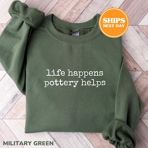 May include: An olive green crewneck sweatshirt featuring the white text "life happens pottery helps" printed on the chest. The garment has long sleeves with ribbed cuffs and a ribbed neckline, constructed from a soft, solid-colored fabric.