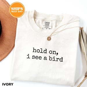 a white t-shirt with the text "hold on, i see a bird" printed on it, along with a necklace and a brown hat.