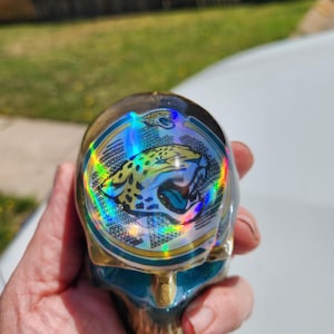 May include: A skull-shaped decorative item with a gold and teal finish. The top features a holographic image of a jaguar with teal and yellow accents. The skull has a metallic sheen and is held in a hand.
