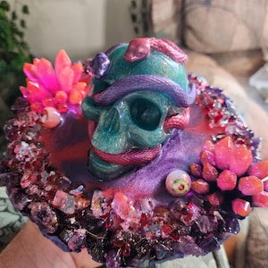 May include: A teal skull sculpture with a purple snake wrapped around it, set on a purple and red base. The base is encrusted with crystals and features pink and red crystal formations. An eyeball is also visible.