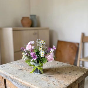 May include: A small, galvanized metal bucket filled with a variety of artificial flowers, including pink roses, white daisies, and purple blooms. The bucket is tied with a green velvet ribbon and sits on a weathered wooden table.