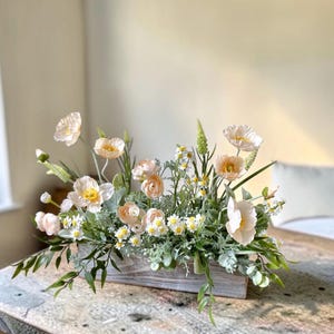 May include: A floral arrangement featuring pale peach poppies, small white daisies, and green foliage in a rectangular, weathered gray wooden planter box. The display includes various stems of greenery and delicate buds arranged horizontally.