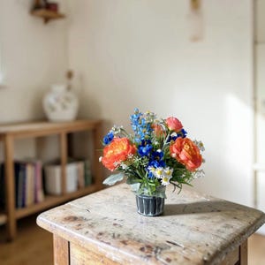 May include: A vibrant floral arrangement in a small, black and white striped pot. The bouquet features orange peonies, blue delphiniums, white daisies, and green foliage. The arrangement sits on a rustic wooden table, creating a charming display.