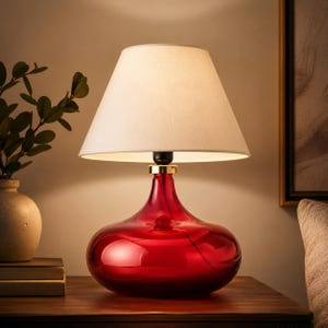 Puede incluir: Una lámpara de mesa con una base ancha de vidrio rojo translúcido de forma lisa y redondeada. La lámpara está rematada con una pantalla cónica de tela color blanco roto. Un pequeño anillo metálico dorado conecta la base con el casquillo.