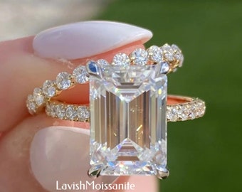 Puede incluir: Un anillo de compromiso de oro con una gran piedra central de corte esmeralda y una banda a juego de diamantes más pequeños. El anillo está engastado en un estilo solitario clásico y está sostenido por una mano con esmalte de uñas blanco. El texto "Lavish Moissanite" es visible en la imagen.