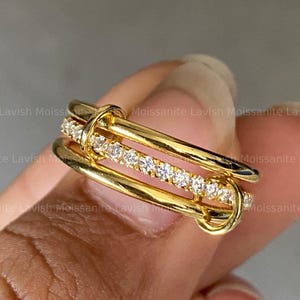 Op de afbeelding: Een goudkleurige ring met een uniek ontwerp. De ring heeft drie parallelle banden, waarvan één is versierd met kleine, sprankelende stenen. De banden zijn verbonden door gebogen, goudkleurige elementen, wat een elegante en moderne uitstraling creëert.