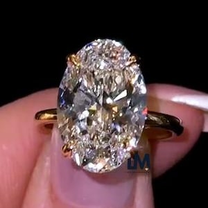 May include: A gold ring featuring a large, oval-cut diamond. The diamond is clear with internal facets, and the ring has a simple, elegant band. The ring is held against a black background.