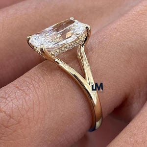 Op de afbeelding: Een gouden ring met een grote, rechthoekig geslepen diamant. De heldere diamant schittert, gezet in een gouden band met kleinere accentstenen. De ring wordt op een vinger getoond, waarbij het ontwerp en vakmanschap worden benadrukt.