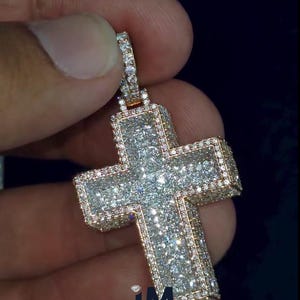 Puede incluir: Un colgante de cruz con incrustaciones de diamantes y un marco dorado. La cruz está cubierta de pequeños diamantes brillantes, con una fianza dorada para sujetarla a una cadena. El colgante tiene el texto "IM LAVISH JEWELRY".